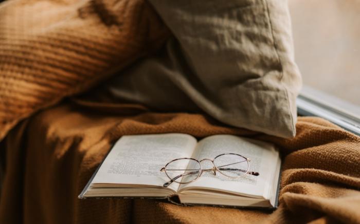 An open book on a windowsill. The book is resting on a knit blanket, and there is a pair of reading glasses on top of the book.